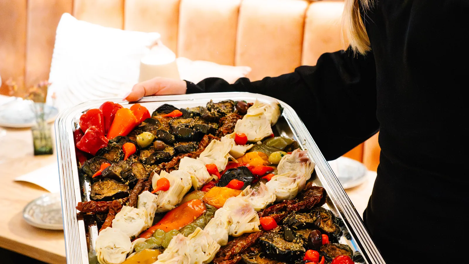 Ein Blech mit gegrillten Artischocken, Paprika, Auberginen und Oliven wird in den Raum getragen