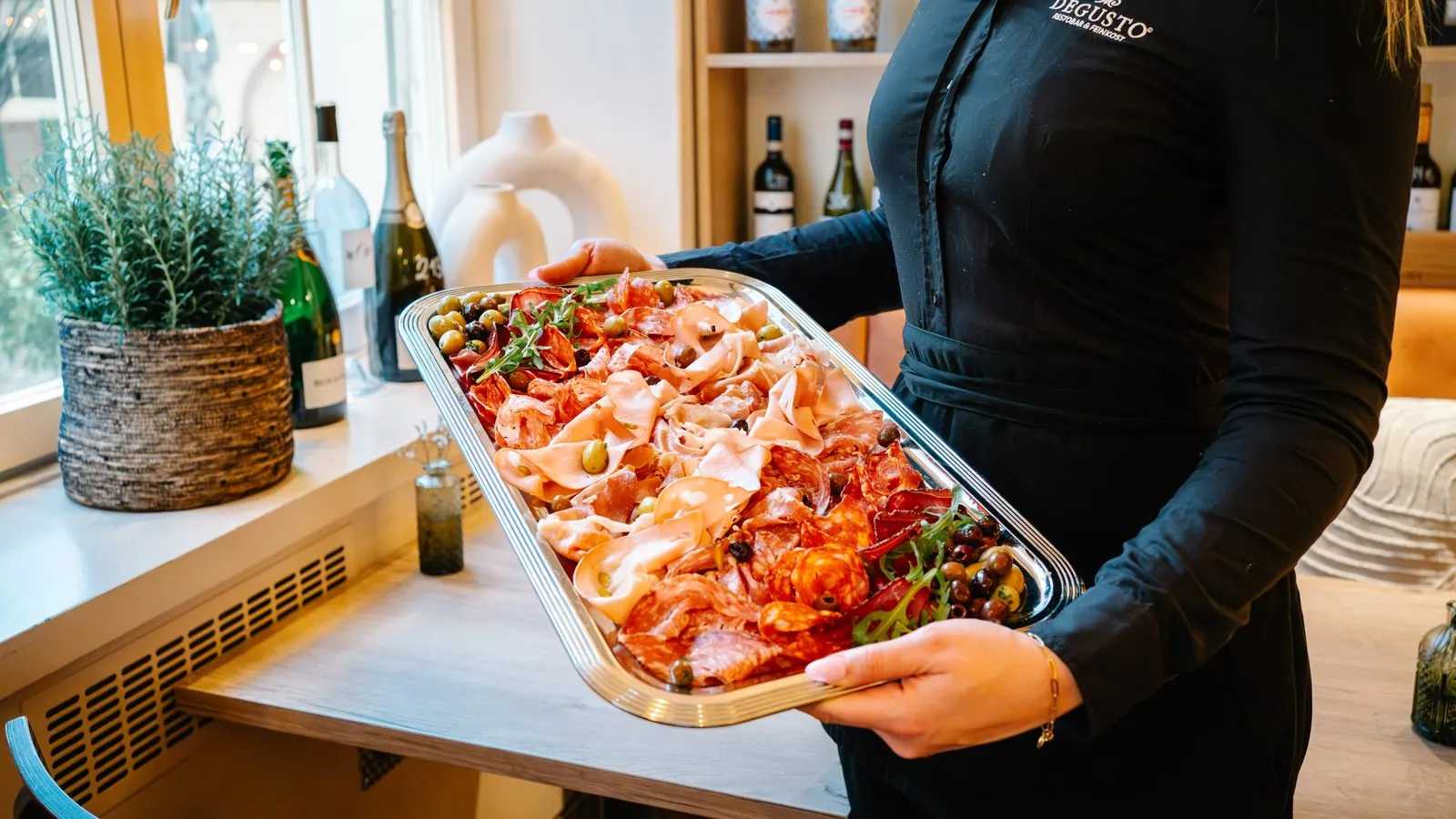 Eine Platte mit Parmaschinken, Salami und Oliven, getragen von einer DEGUSTO-Mitarbeiterin