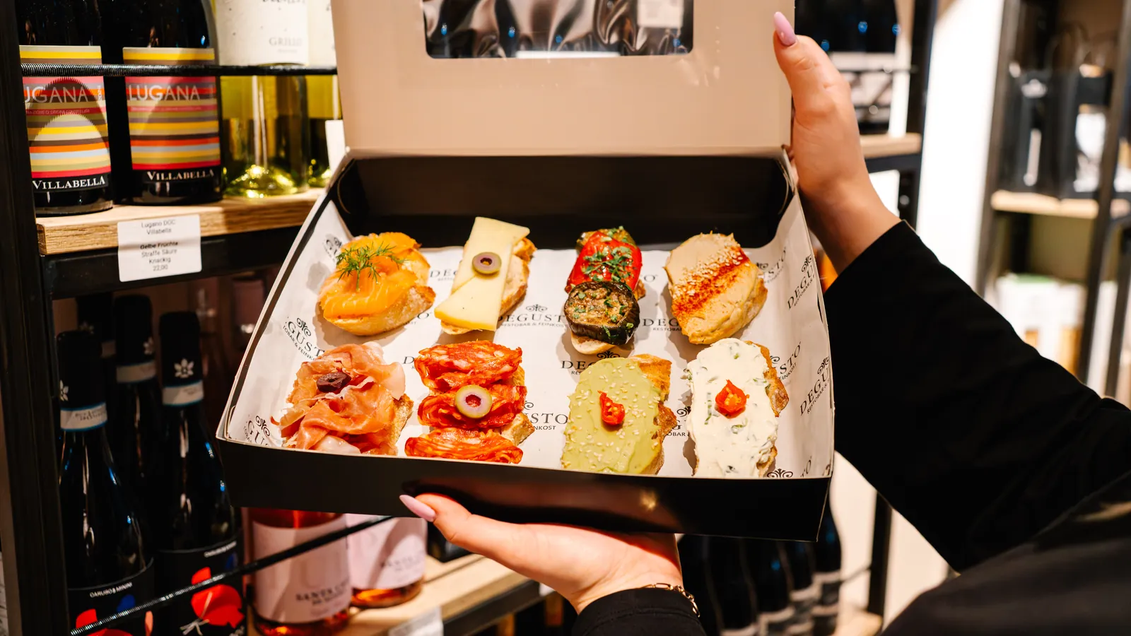 Eine geöffnete Box mit belegten Canapés vor dem Weinregal