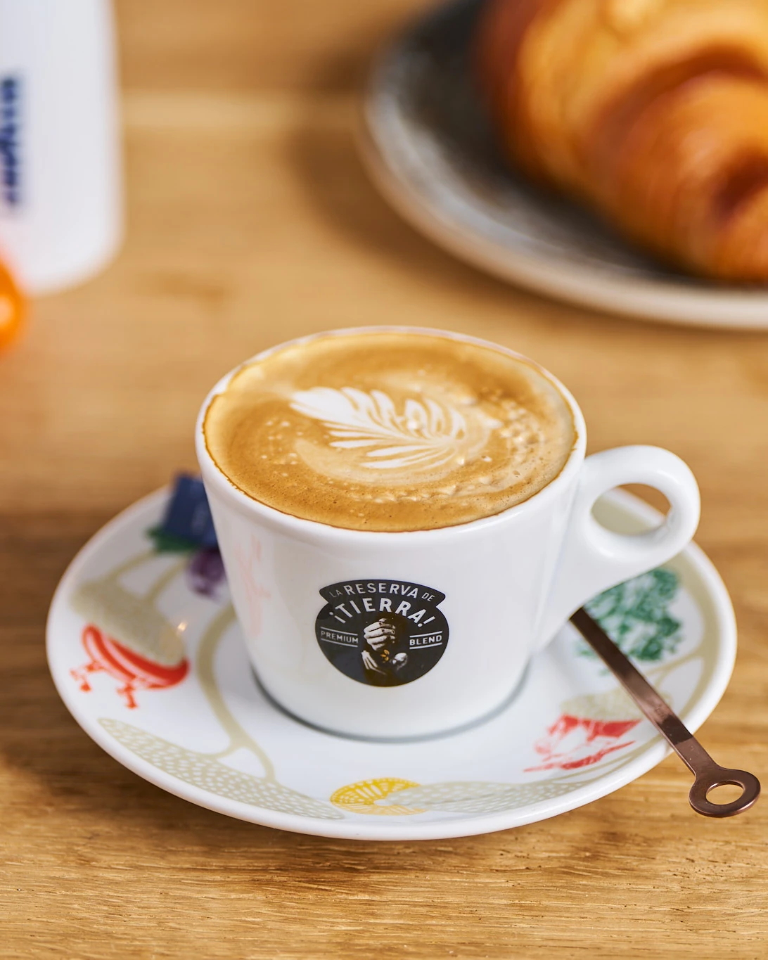 Cappuccino mit Milchschaum-Muster in einer DEGUSTO-Tasse, dahinter ein Croissant und ein Lavazza-Kännchen