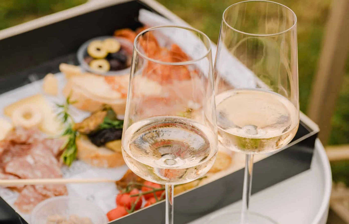 Eine geöffnete Cateringbox mit Schinken, Käse, Oliven und Brot, davor zwei Gläser Weißwein