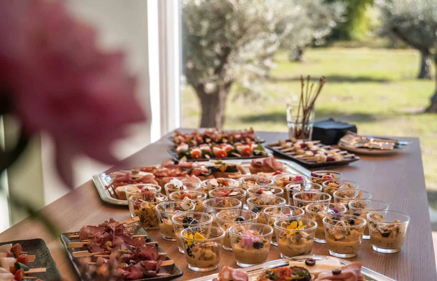 Eine lange Tafel mit Salatgläsern, Caprese-Spießen und Schinkenplatten, dahinter der Blick durch die Fensterfront auf Olivenbäume