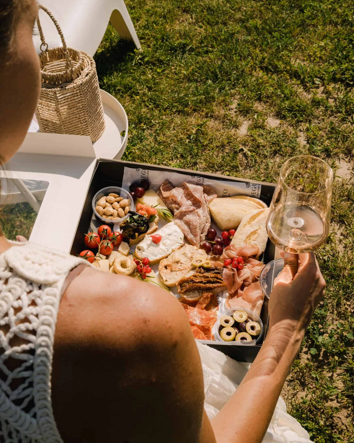 Eine geöffnete Cateringbox mit Schinken, Käse, Tomaten und Nüssen auf dem Schoß, daneben ein Glas Wein, draußen auf der Wiese