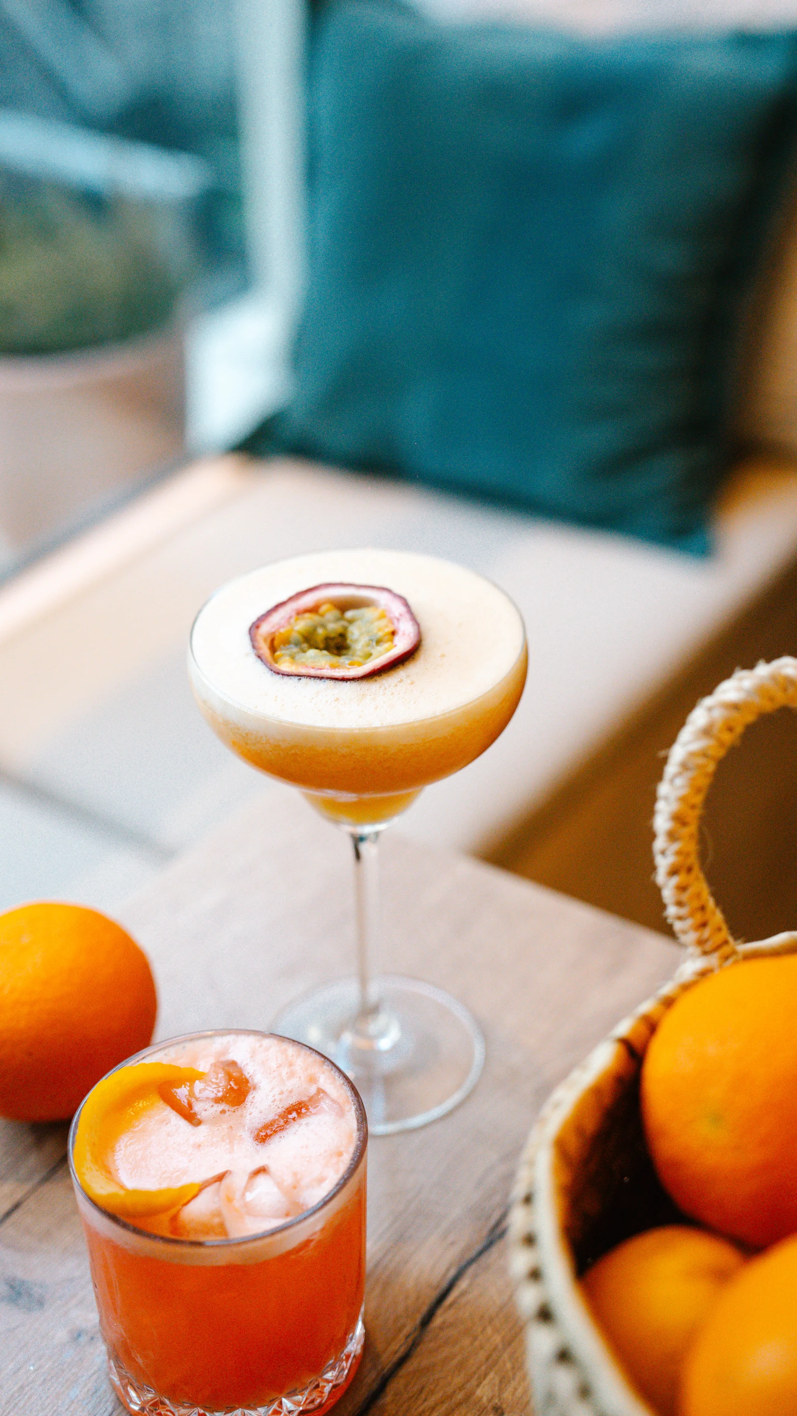 Ein Cocktail im Coupé-Glas mit halber Maracuja auf dem Schaum, daneben ein Aperitif auf Eis und ein Korb mit Orangen