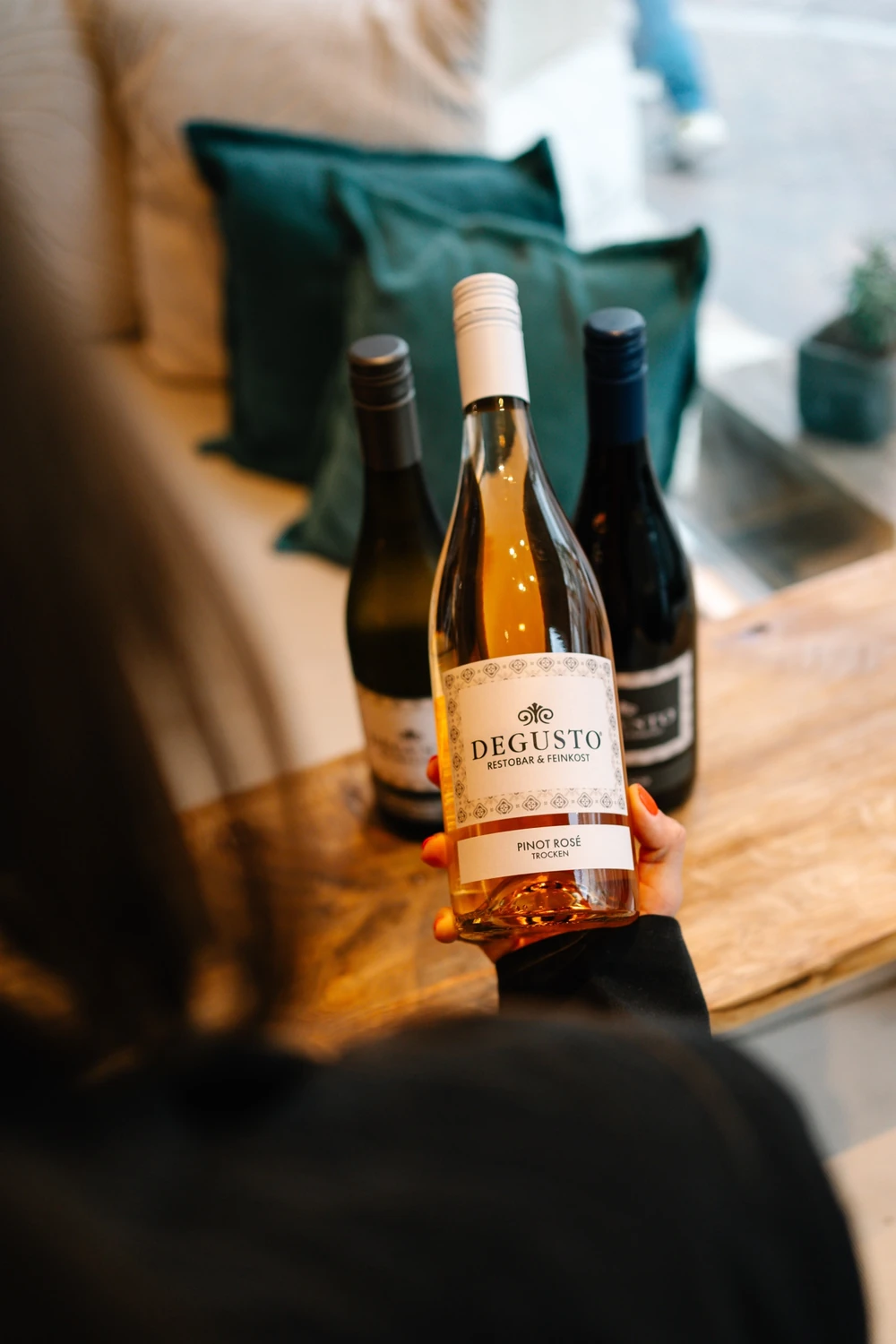 Eine Flasche DEGUSTO Pinot Rosé wird am Fenstertisch in der Hand gehalten, dahinter zwei weitere Eigenweine