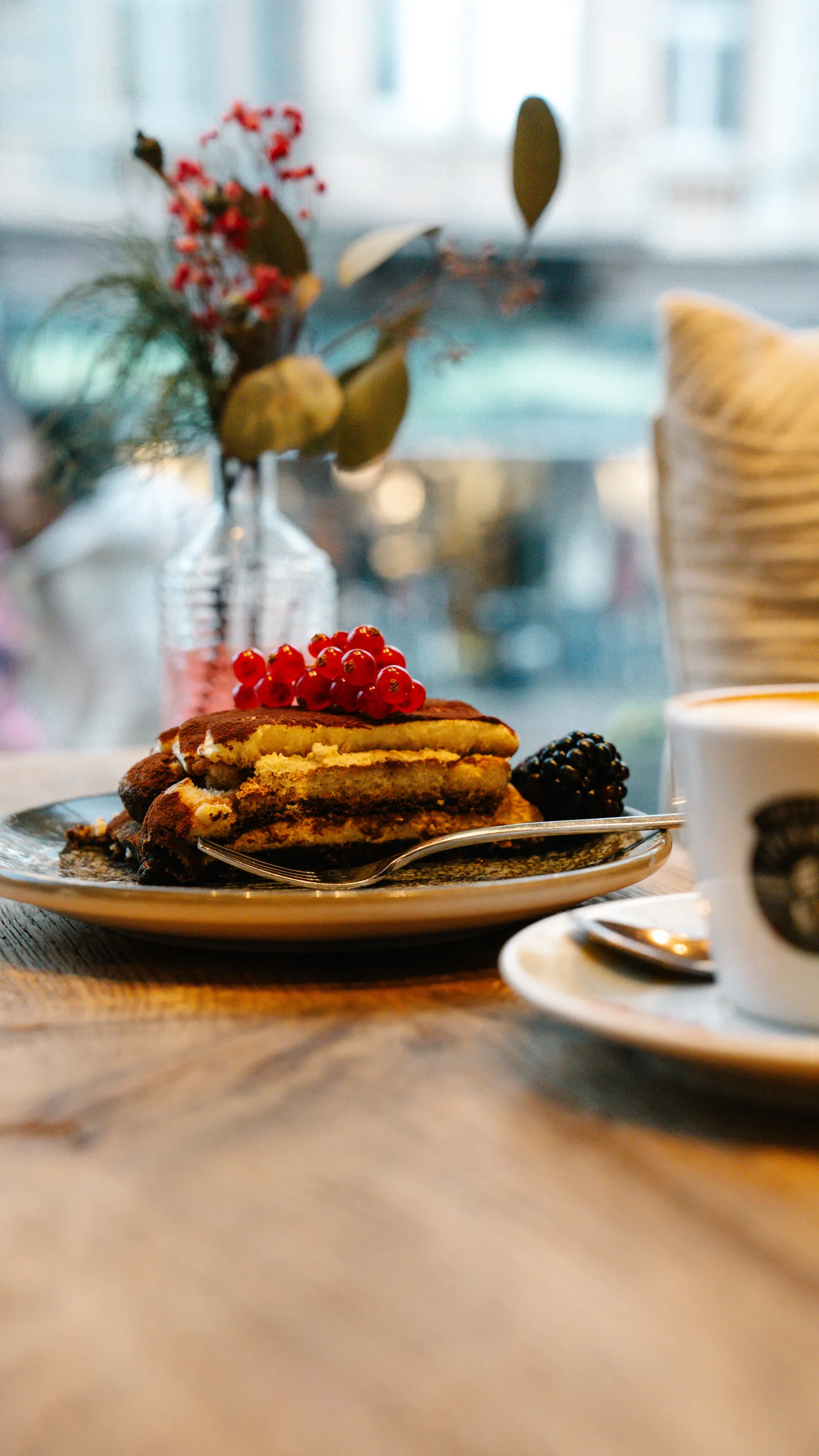 Tiramisu mit roten Johannisbeeren und einer Brombeere, daneben eine Tasse Cappuccino am Fenstertisch