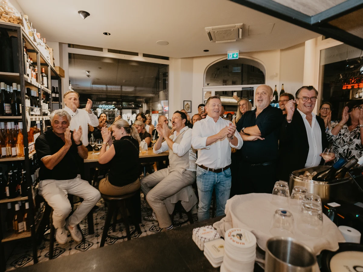 Eine Gästerunde im Gastraum des DEGUSTO applaudiert, dahinter das Weinregal