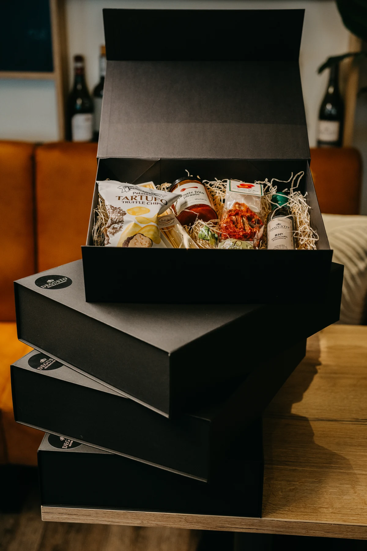 Vier schwarze Genussboxen übereinander gestapelt, die oberste geöffnet mit Trüffelchips, Sugo, getrockneten Tomaten und Olivenöl in Holzwolle