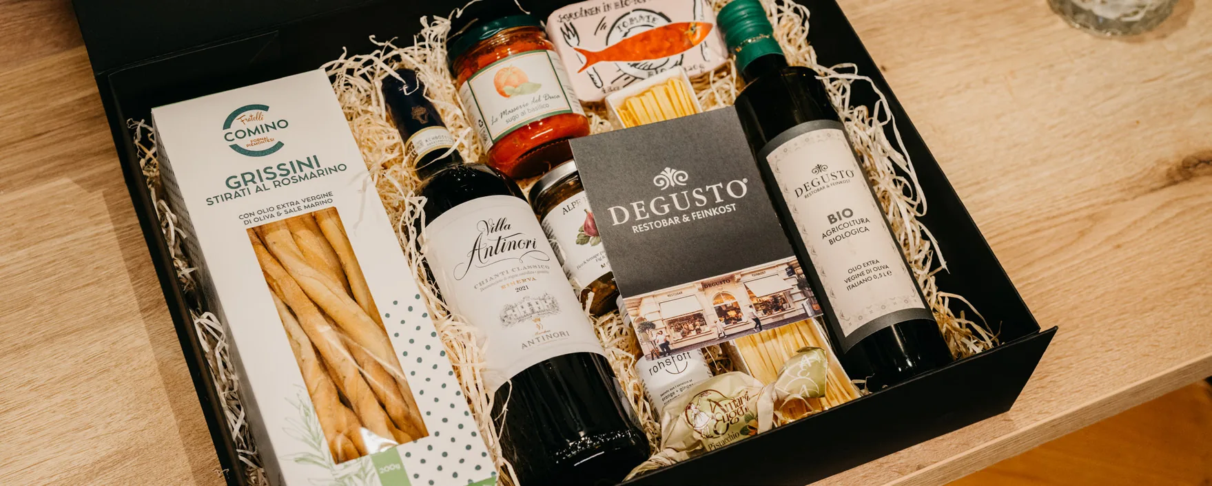 Eine offene Geschenkbox in Holzwolle mit Grissini, Sugo, Olivenöl, Pasta und einer Flasche Wein