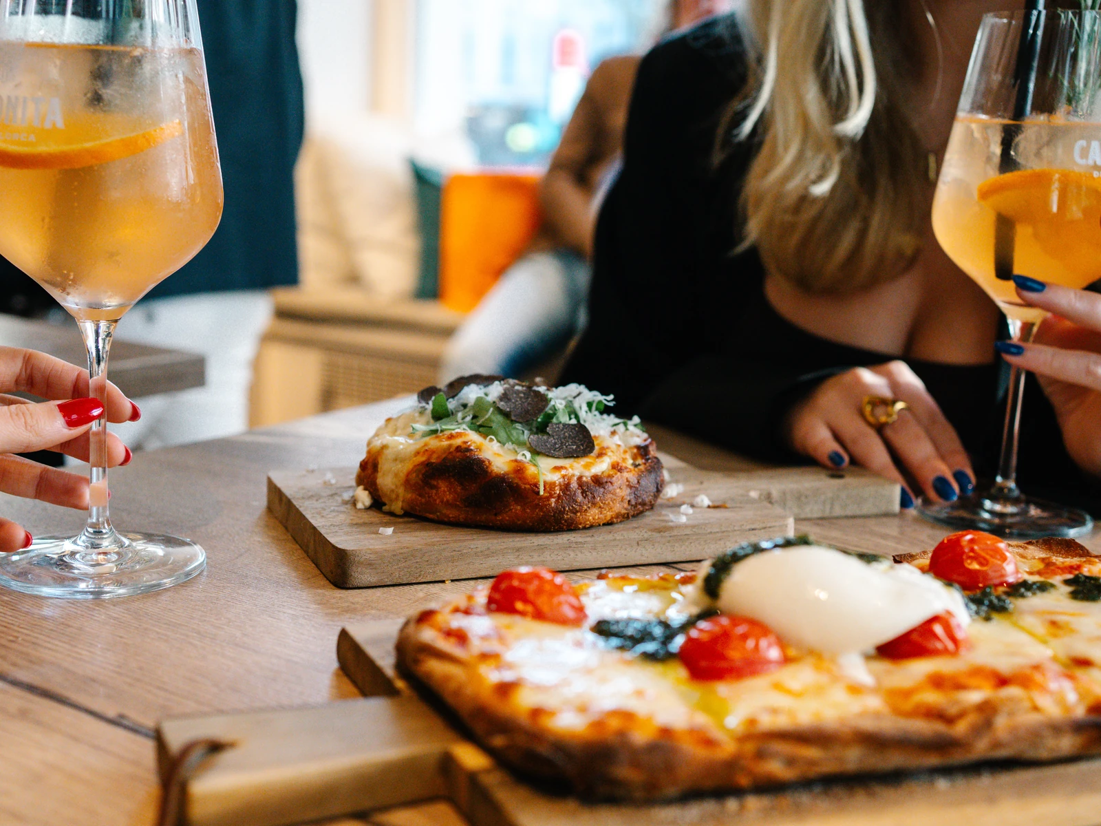 Zwei Pinsa auf Holzbrettern, eine mit Trüffel, eine mit Tomaten und Burrata, dahinter zwei Gäste mit Aperitivo