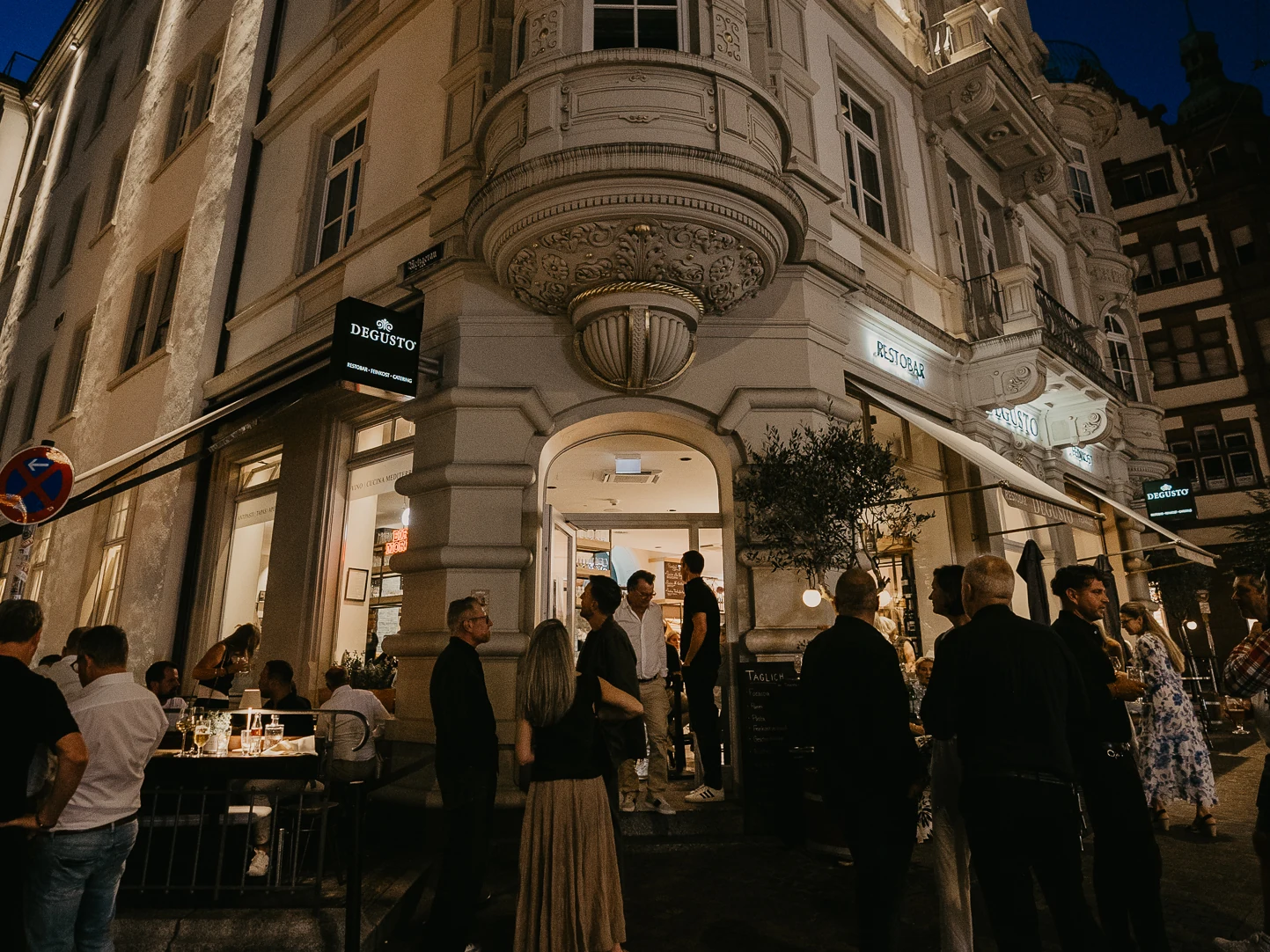 Die Fassade des DEGUSTO in der Dämmerung, davor Gäste auf der Straße