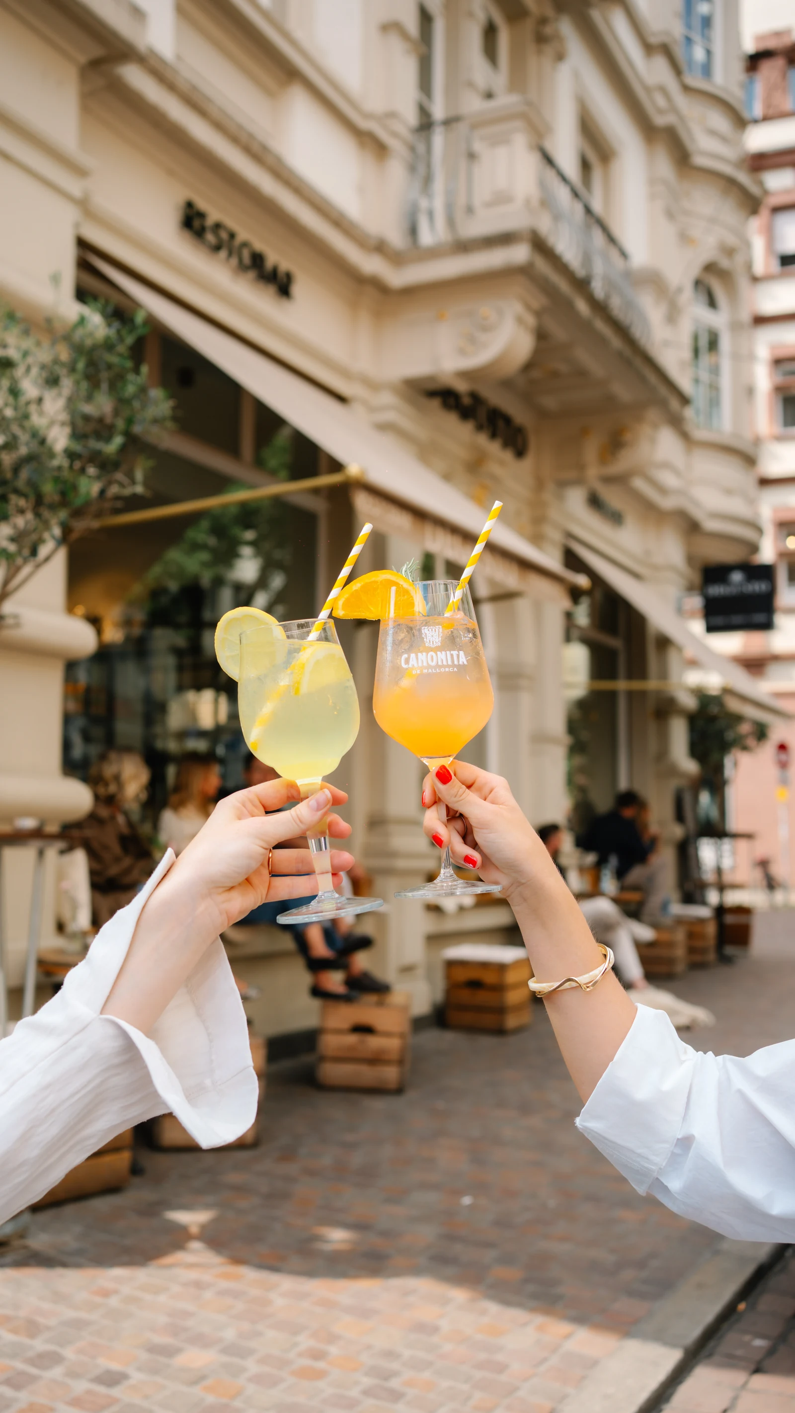 Zwei Hände stoßen mit Spritz-Gläsern an, im Hintergrund die Fassade des DEGUSTO mit Markise