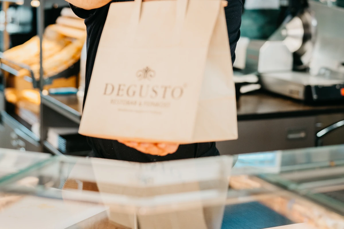 Ein Mitarbeiter reicht eine gefüllte DEGUSTO-Papiertüte über die Theke