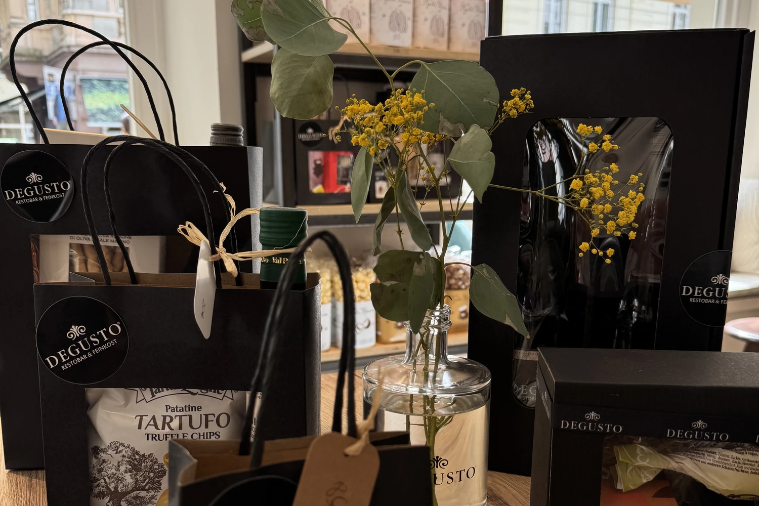 Mehrere DEGUSTO-Geschenktüten und -boxen mit Sichtfenster auf dem Fensterbrett, daneben ein Strauß